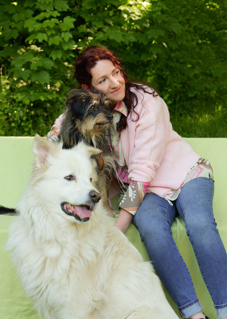 Eine Frau sitzt auf einer Bank, schaut nach rechts und lächelt. Sie trägt Jeans und eine rosa Strickjacke. Neben ihr sitzt ein kleiner, schwarz-brauner zotteliger Hund und darunter, auf dem Boden, sitzt ein großer, weißer Hund. Beide Hunde schauen ebenfalls nach rechts.